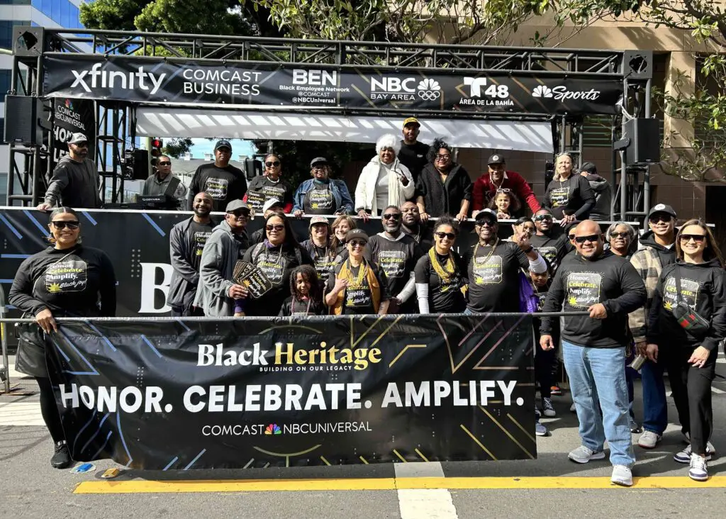 A large group of parade attendees in front of a stage at the 2026 Black Joy Parade. People in the front row are holding a sign that reads 'Honor, Celebrate, Amplify'.